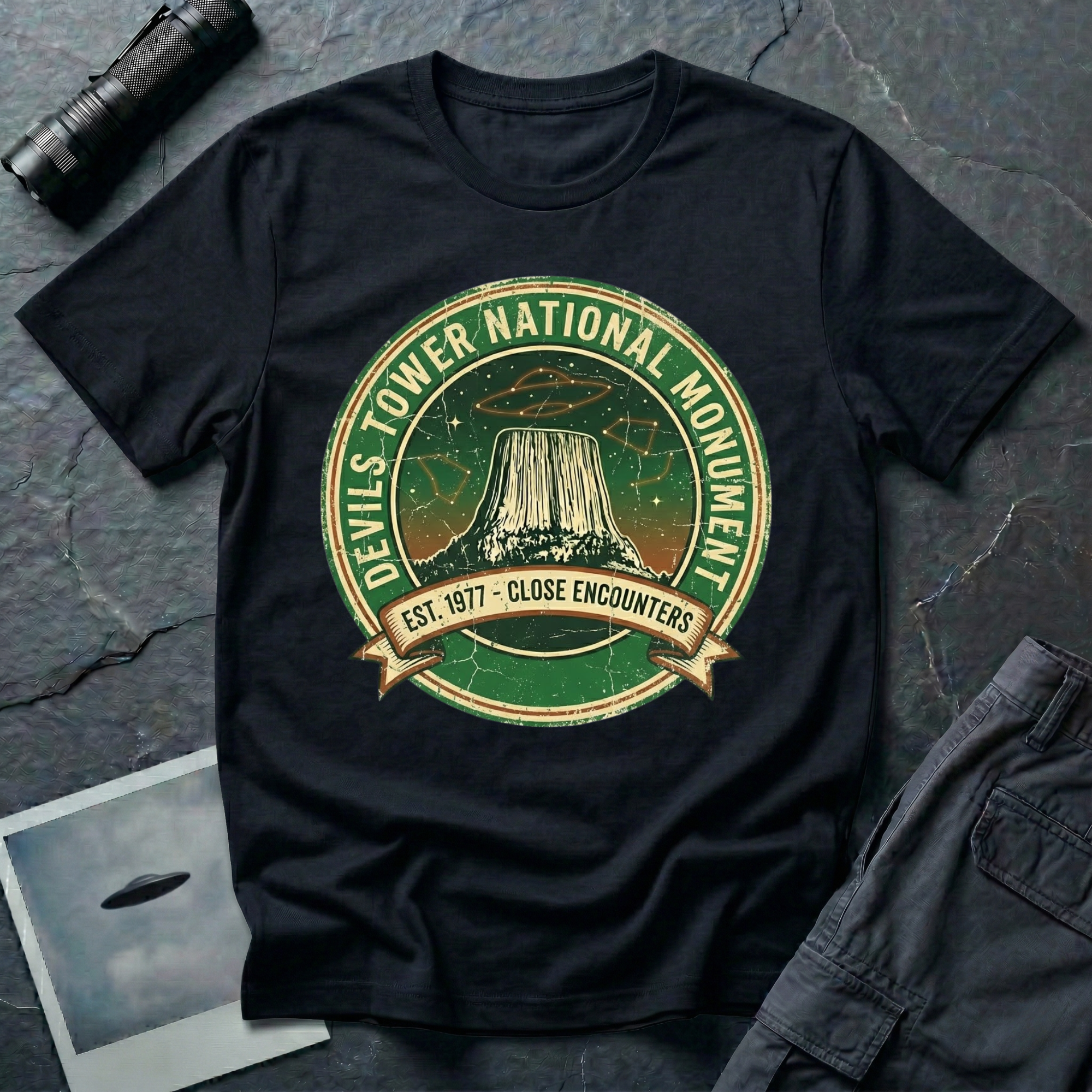 Devils Tower UFO t-shirt — vintage distressed National Park badge with the Wyoming monument silhouette, tiny flying saucers and constellations over a starry green sky, "DEVILS TOWER NATIONAL MONUMENT" arc text and "EST. 1906 — CLOSE ENCOUNTERS" banner on black cotton tee