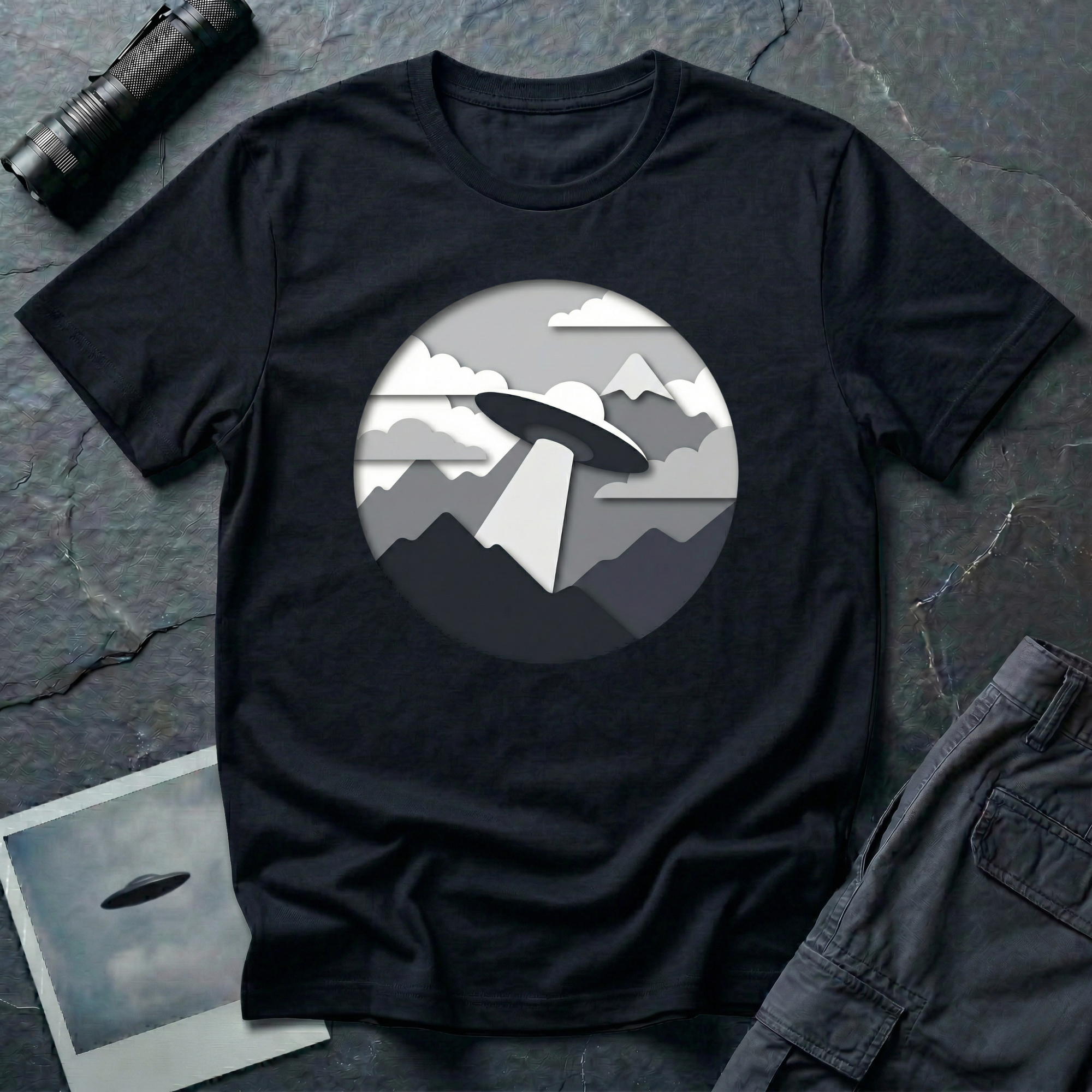 Papercut UFO t-shirt — layered paper-craft illustration of flying saucer over mountains and clouds in grey monochrome on black cotton tee