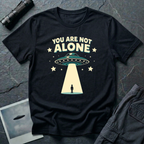 You Are Not Alone friendly UFO t-shirt — retro saucer beaming down on a calm human figure with "You Are Not Alone" text on black cotton tee