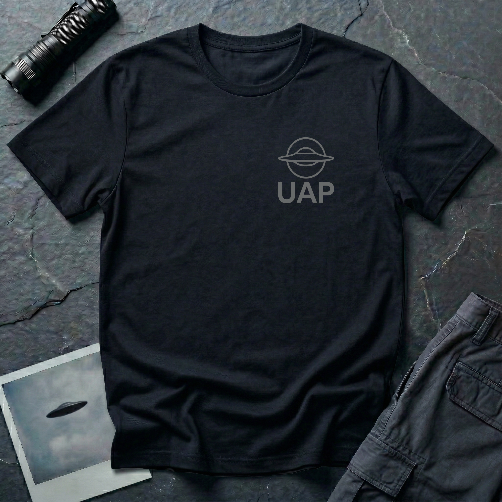 UAP logo t-shirt — small minimalist saucer icon and UAP letters in grey on the upper left chest of black cotton tee