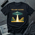 Welcome Visitors Southwest UFO t-shirt — flying saucer beaming down into gold and teal desert canyon landscape with "Welcome Visitors" text on black cotton tee