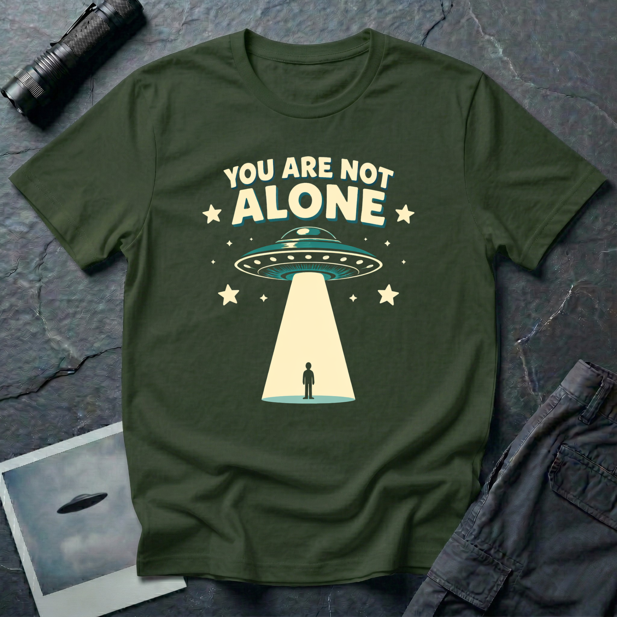 You Are Not Alone friendly UFO t-shirt — retro saucer beaming down on a calm human figure with "You Are Not Alone" text on military green cotton tee