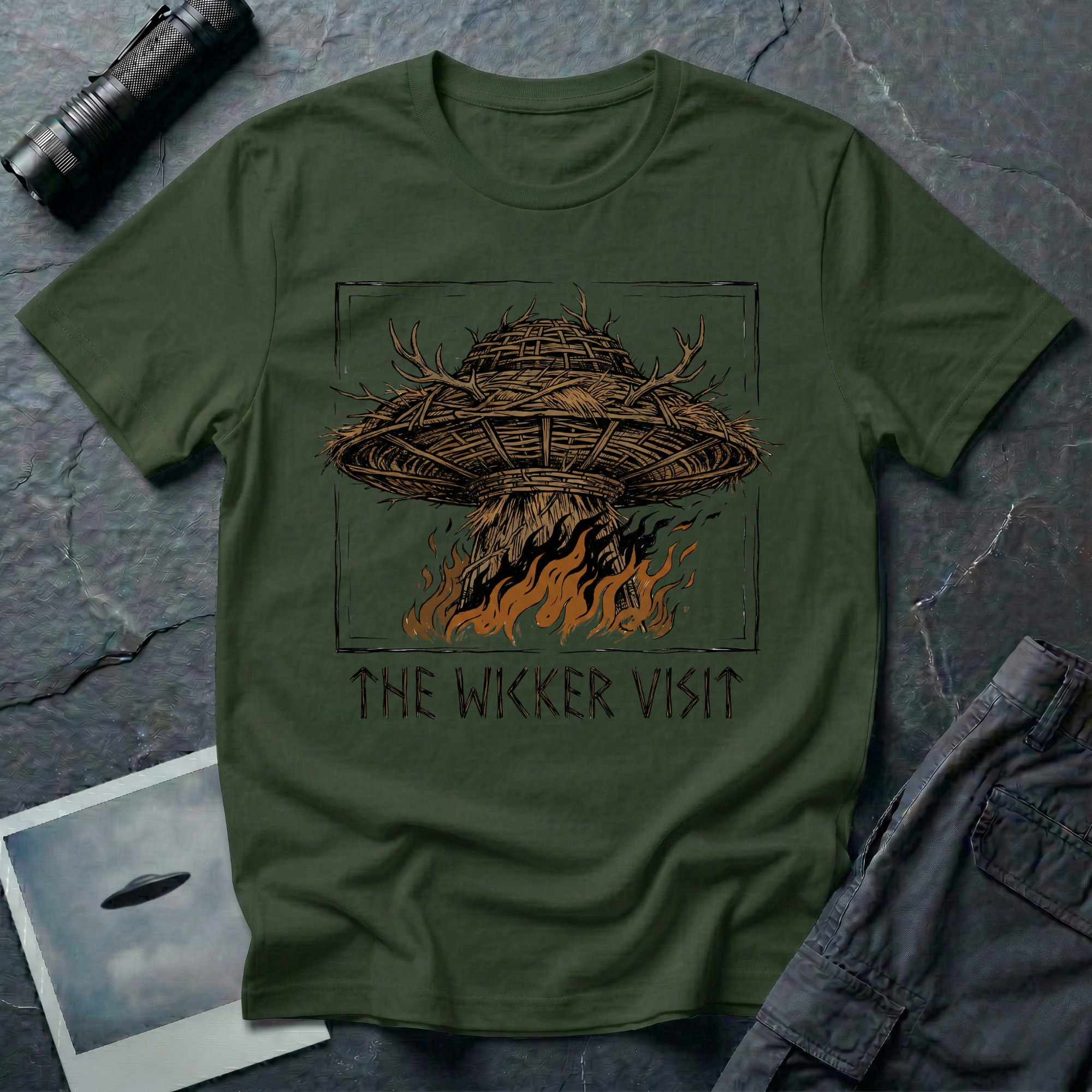 The Wicker Visit folk horror UFO t-shirt — woven wicker flying saucer with antler crown burning in orange flames on military green tee