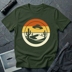 Desert Sun sunset UFO t-shirt — classic flying saucer silhouette over layered retro sunset sky with burnt orange and warm yellow bands, two saguaro cacti and a cream desert dune in a clean circular graphic composition on dark green cotton tee