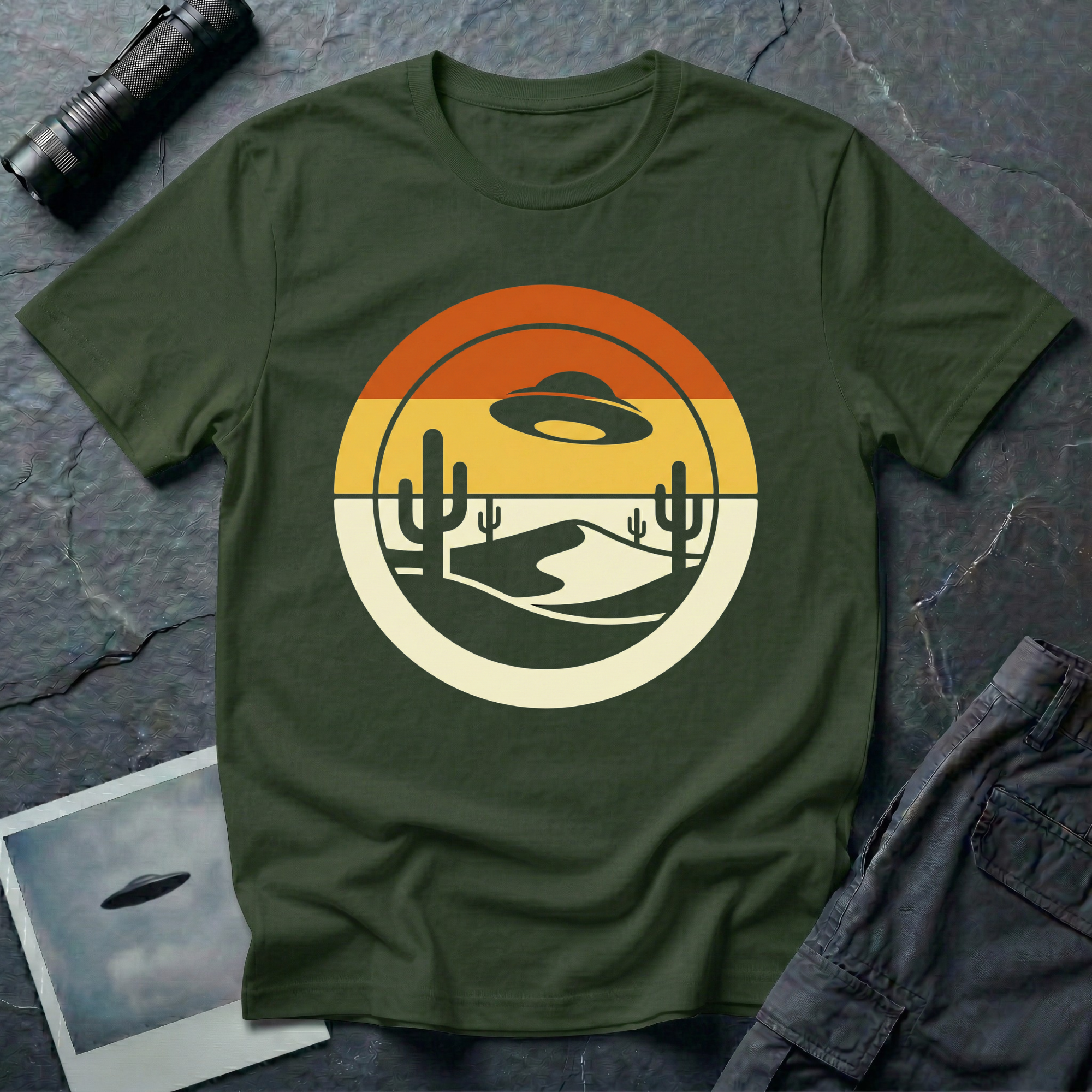 Desert Sun sunset UFO t-shirt — classic flying saucer silhouette over layered retro sunset sky with burnt orange and warm yellow bands, two saguaro cacti and a cream desert dune in a clean circular graphic composition on dark green cotton tee