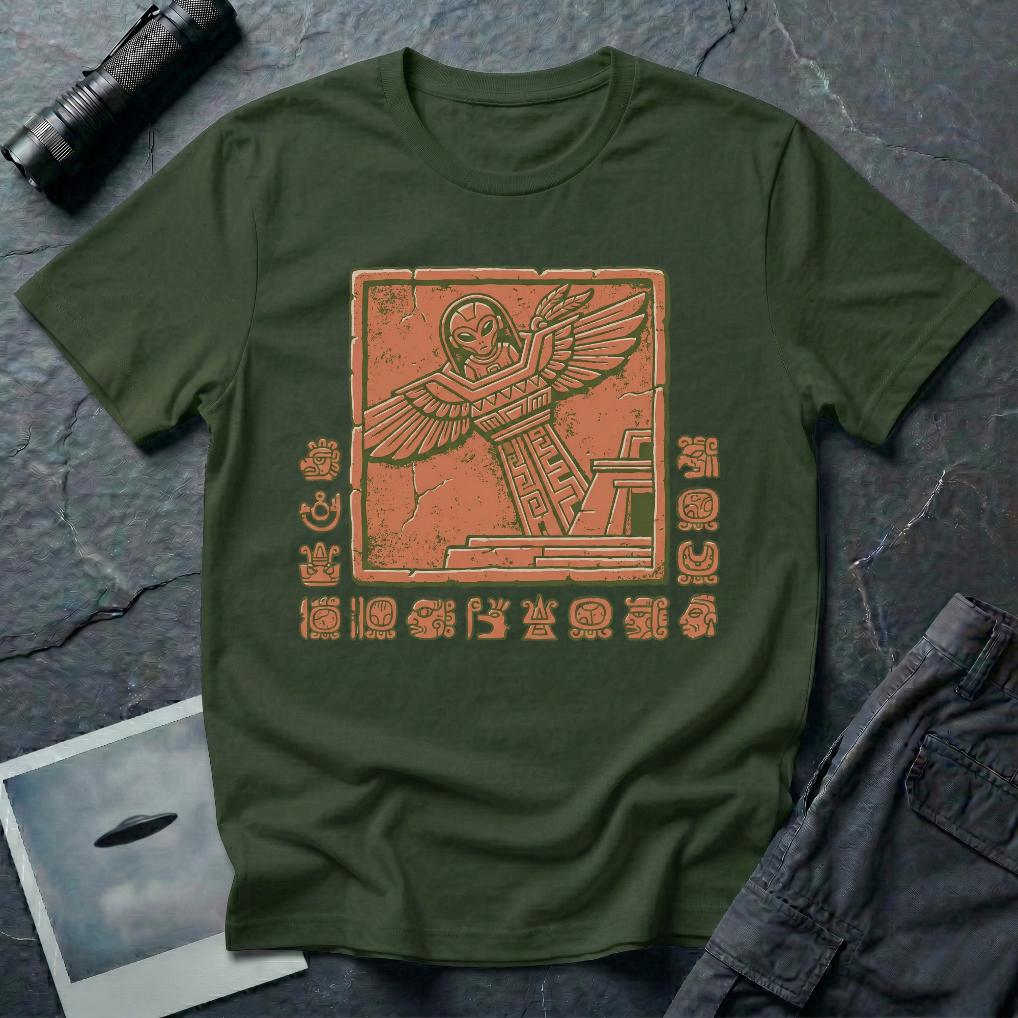 Aztec alien t-shirt — ancient astronaut figure with grey alien head in Mesoamerican feathered dress on stepped pyramid. Weathered stone relief on green cotton tee.