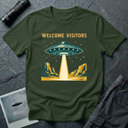 Welcome Visitors Southwest UFO t-shirt — flying saucer beaming down into gold and teal desert canyon landscape with "Welcome Visitors" text on military green cotton tee