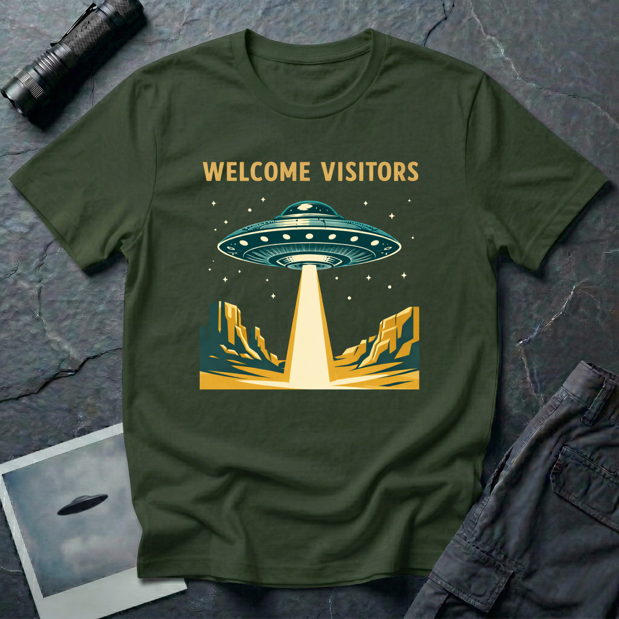 Welcome Visitors Southwest UFO t-shirt — flying saucer beaming down into gold and teal desert canyon landscape with "Welcome Visitors" text on military green cotton tee