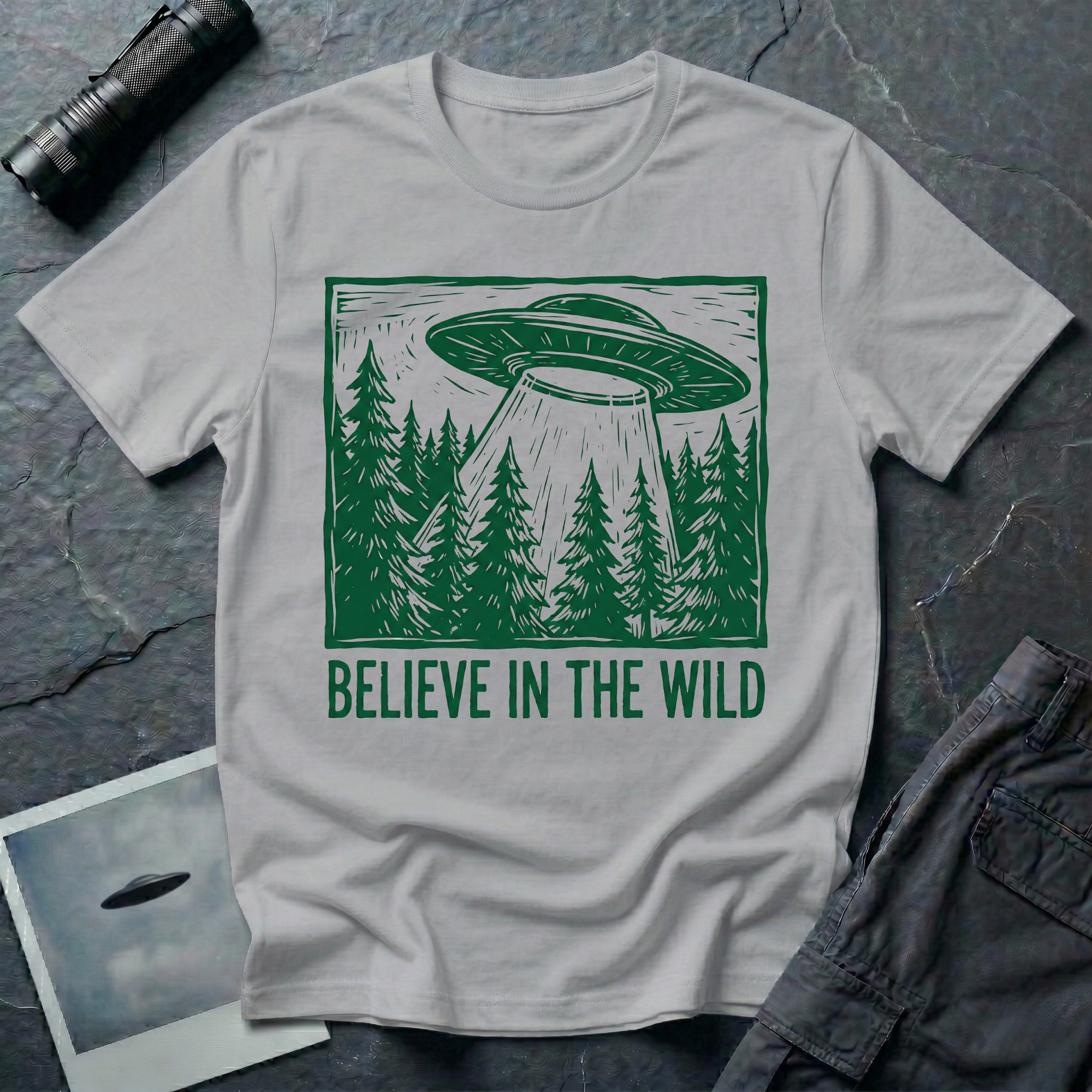 Believe In The Wild forest UFO t-shirt — flying saucer over dense pine forest rendered in green woodcut linocut carved-block print style with "BELIEVE IN THE WILD" caption on grey cotton tee