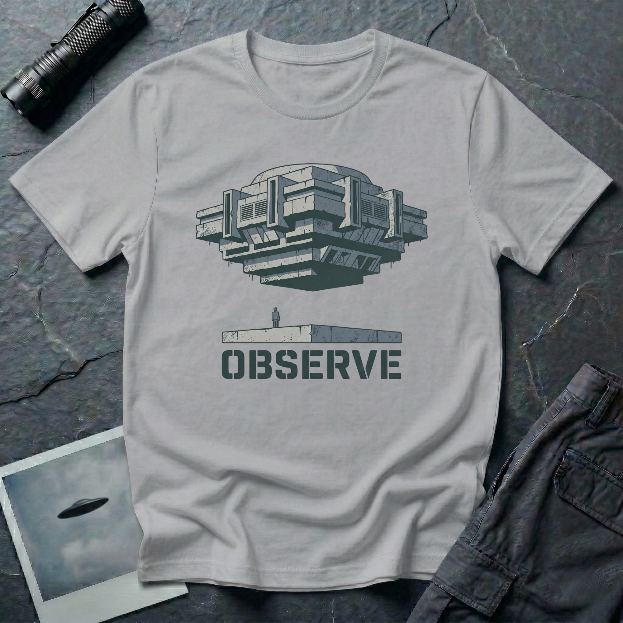 Observe Brutalist UFO t-shirt — massive concrete architectural craft hovering over small figure on plinth with "Observe" stencil type on grey cotton tee