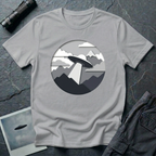 Papercut UFO t-shirt — layered paper-craft illustration of flying saucer over mountains and clouds in grey monochrome on grey cotton tee