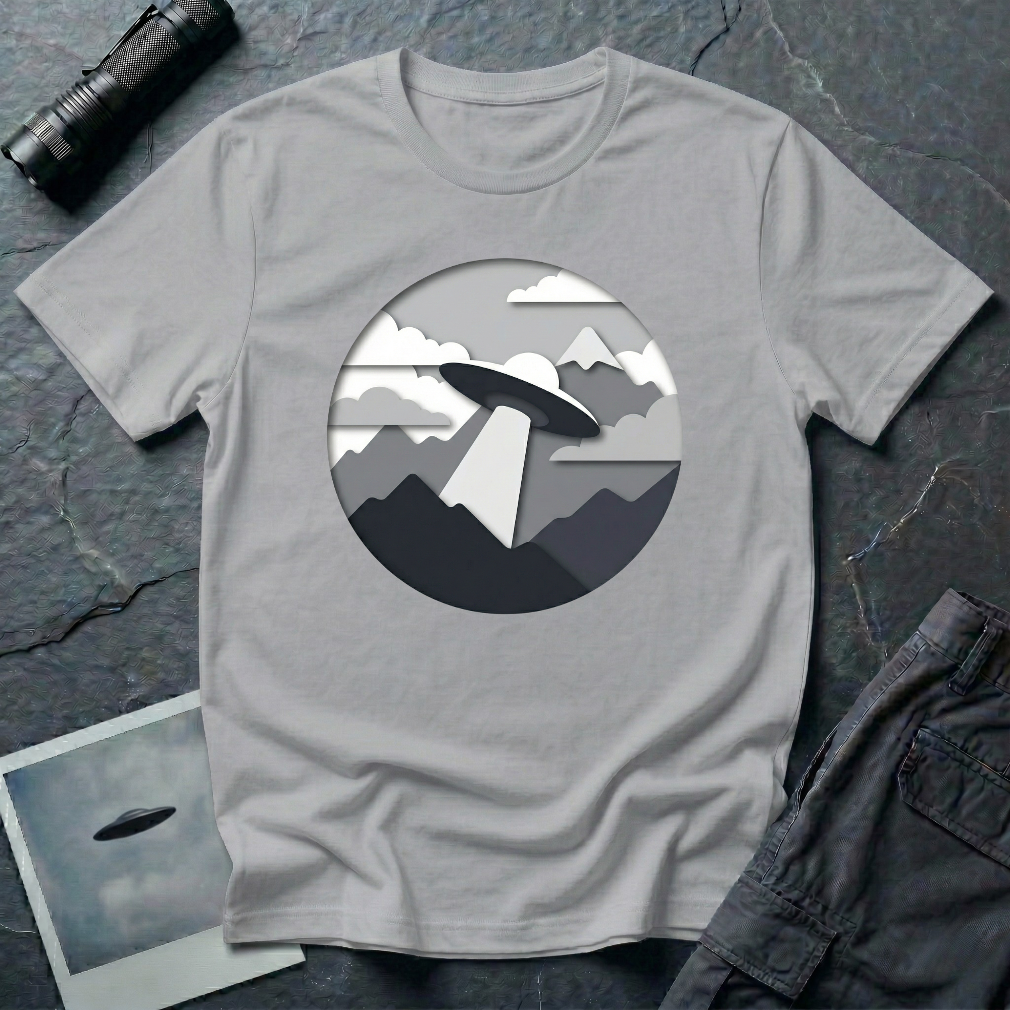 Papercut UFO t-shirt — layered paper-craft illustration of flying saucer over mountains and clouds in grey monochrome on grey cotton tee