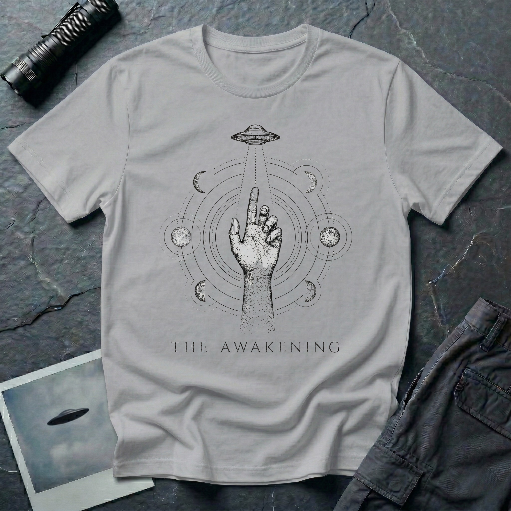 The Awakening mystical UFO t-shirt — hand reaching toward flying saucer beam surrounded by six moon phases in stippled black dotwork on grey cotton tee