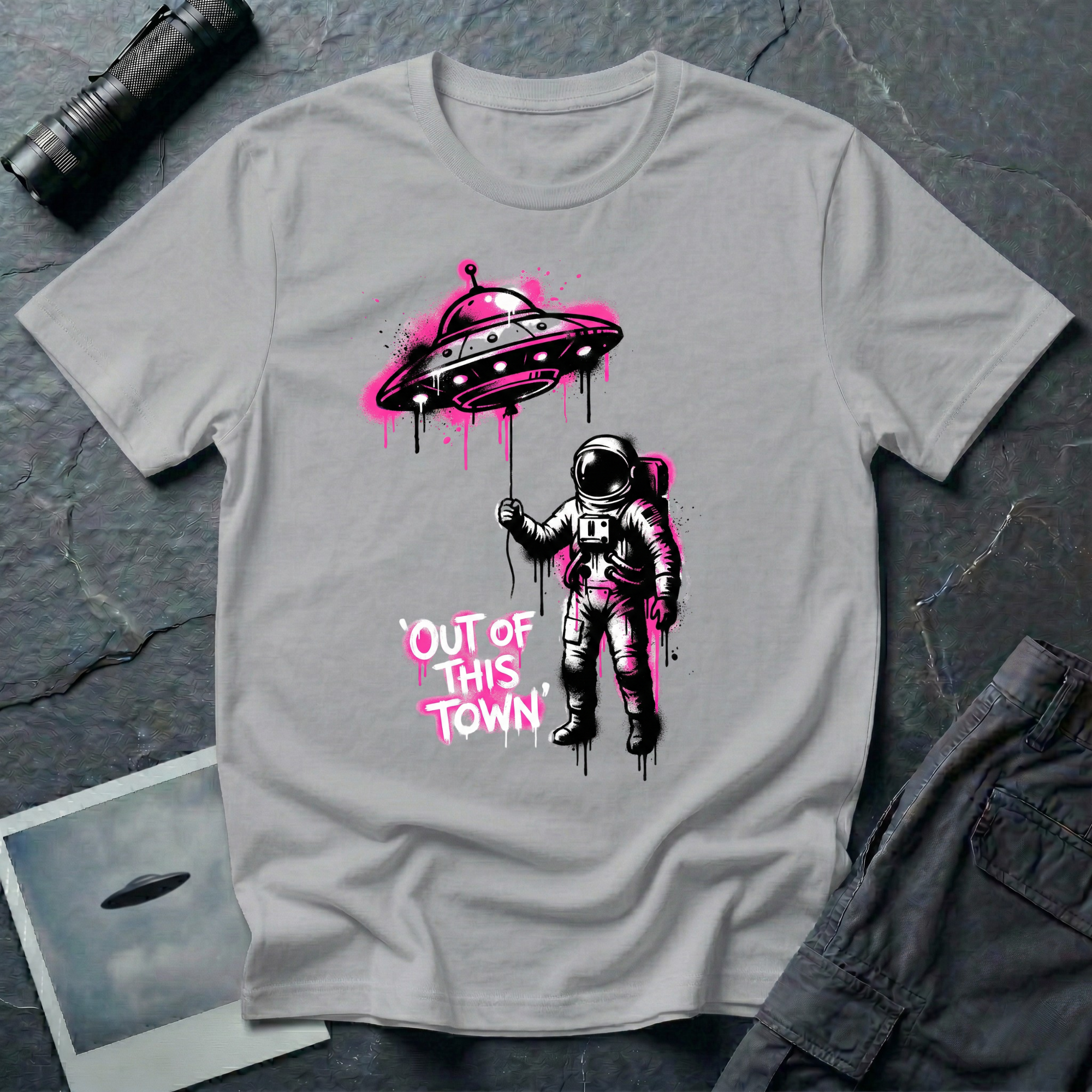 Out of This Town stencil UFO t-shirt — astronaut holding flying saucer like a balloon in Banksy-style street art with hot pink accents on grey cotton tee