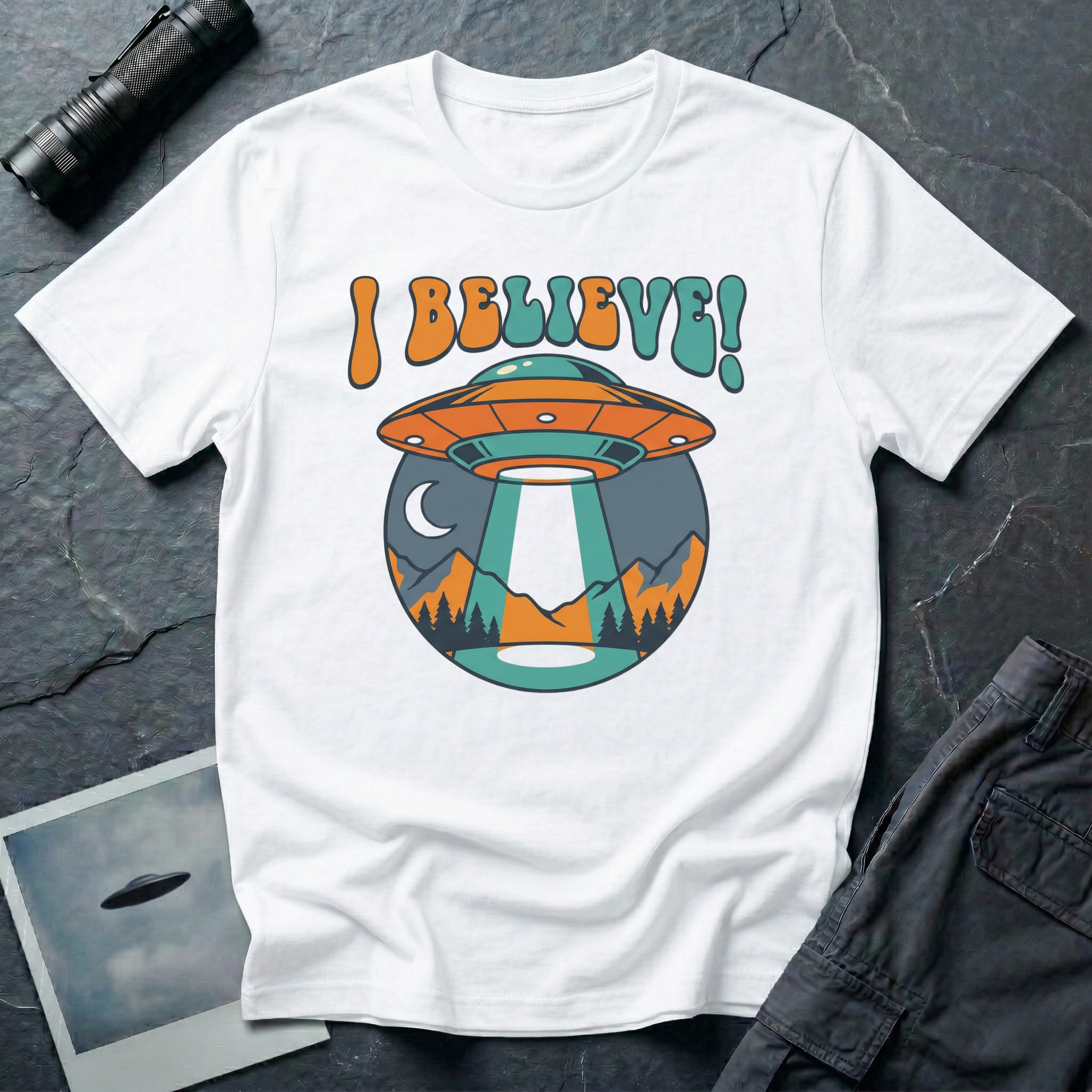 I Believe retro 70s UFO t-shirt — groovy flying saucer beaming down on moonlit mountain scene with bubble typography in orange and teal on white cotton tee