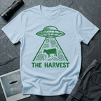 The Harvest dark folklore UFO t-shirt — linocut flying saucer with swirling beam abducting a lone cow in green on light blue cotton tee