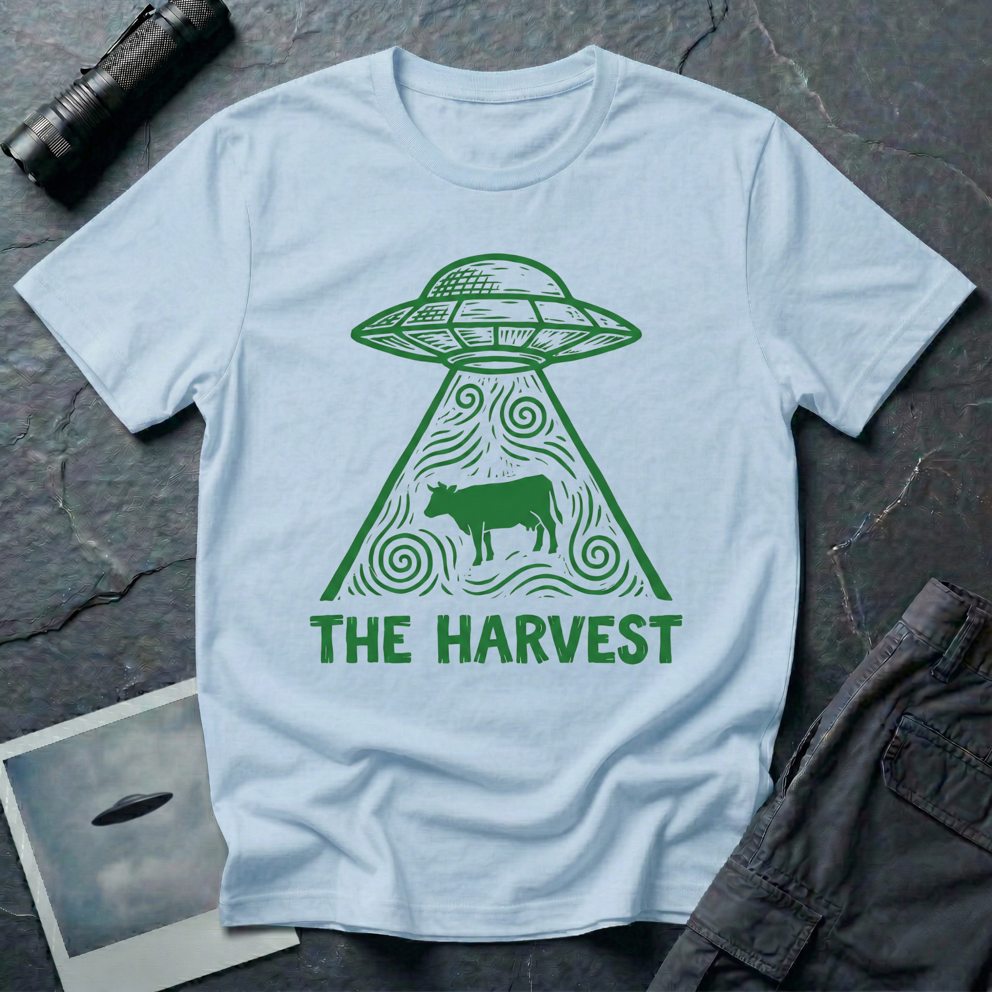 The Harvest dark folklore UFO t-shirt — linocut flying saucer with swirling beam abducting a lone cow in green on light blue cotton tee