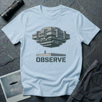 Observe Brutalist UFO t-shirt — massive concrete architectural craft hovering over small figure on plinth with "Observe" stencil type on pale blue cotton tee
