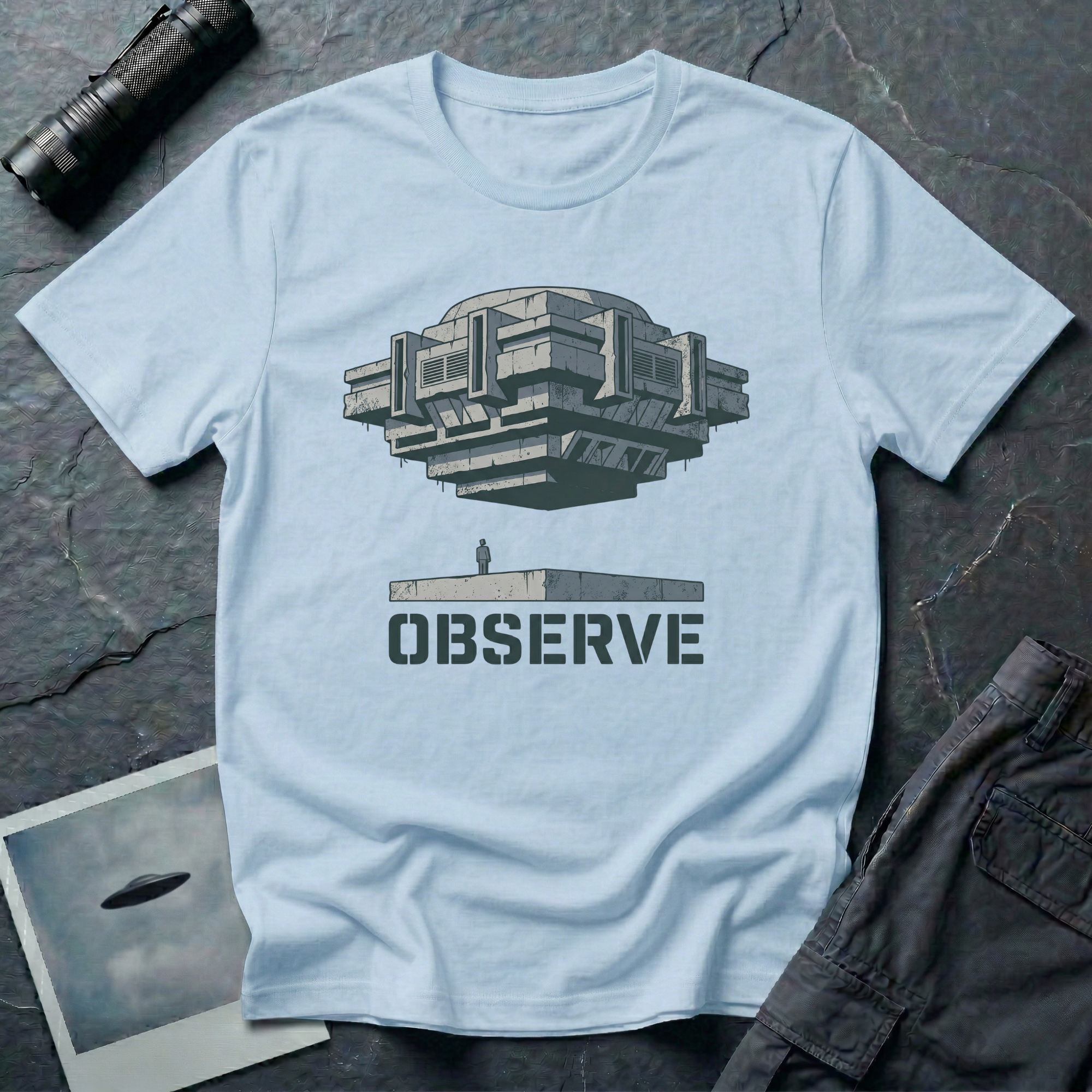 Observe Brutalist UFO t-shirt — massive concrete architectural craft hovering over small figure on plinth with "Observe" stencil type on pale blue cotton tee