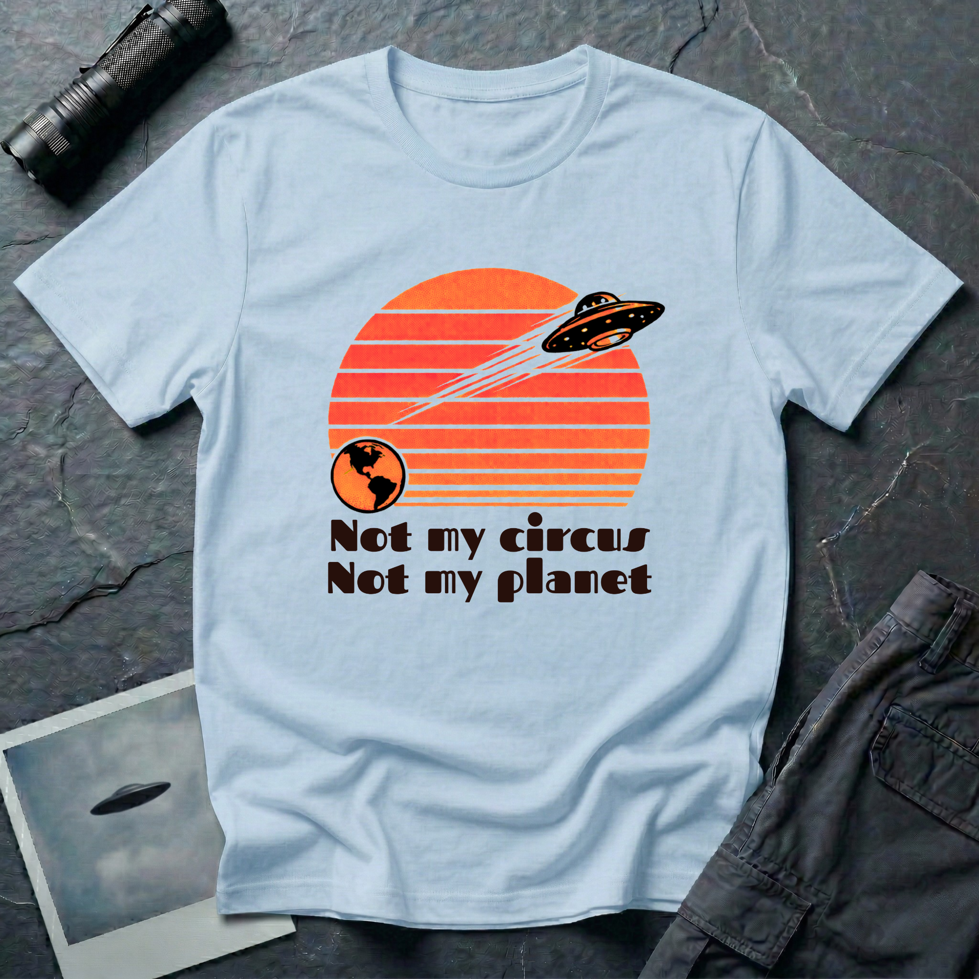 Not My Circus retro 1970s UFO t-shirt — flying saucer leaving Earth against striped sunset with "not my circus, not my planet" type on light blue unisex cotton tee