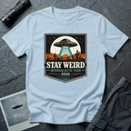 Stay Weird desert UFO t-shirt — tour-poster badge with flying saucer over desert landscape and cacti on light blue cotton tee
