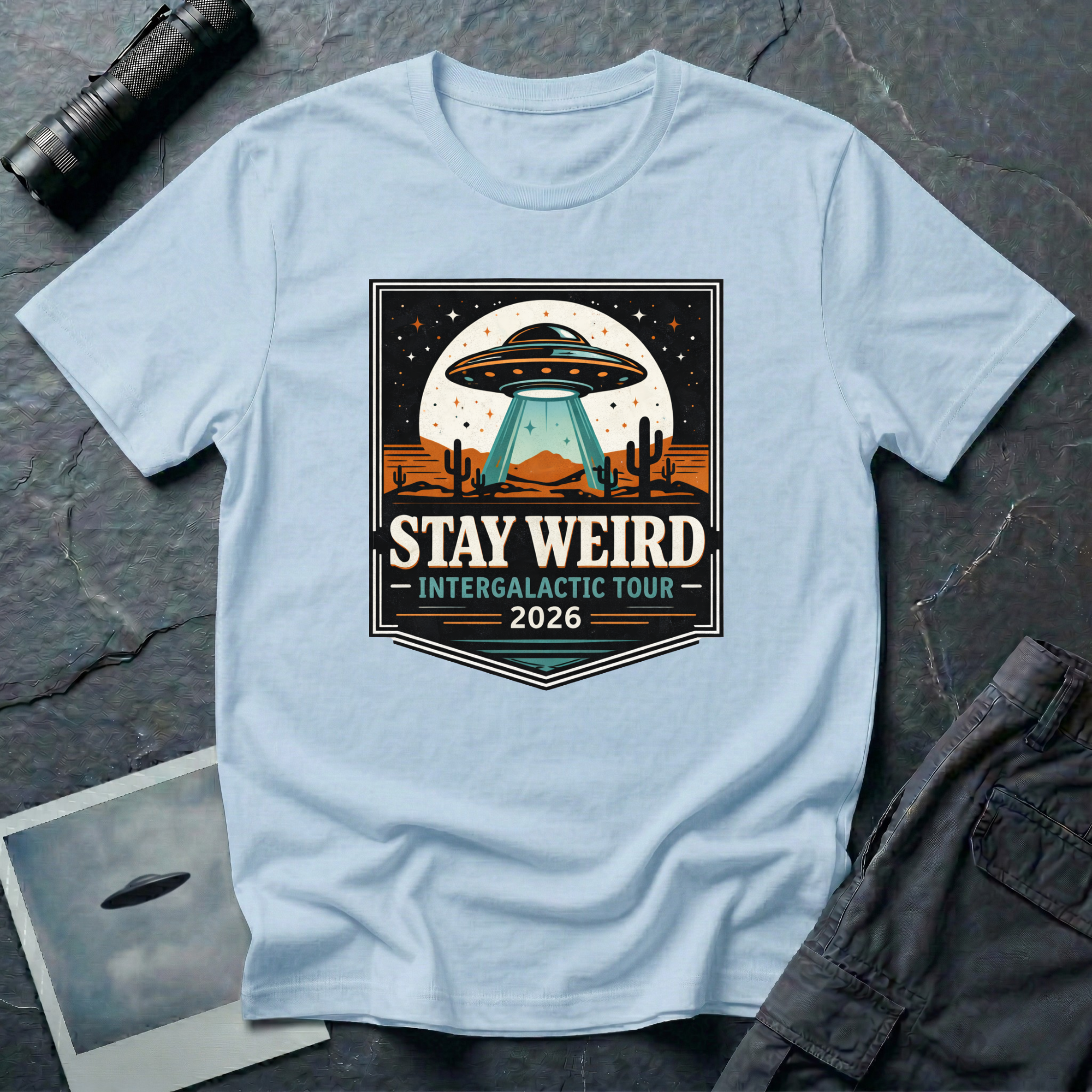 Stay Weird desert UFO t-shirt — tour-poster badge with flying saucer over desert landscape and cacti on light blue cotton tee