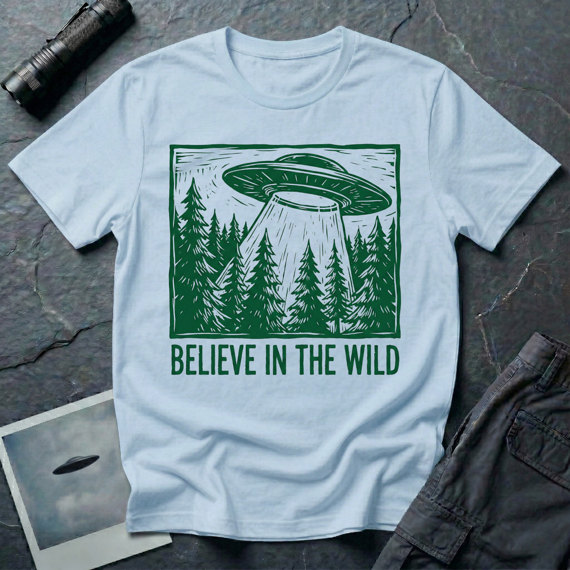 Believe In The Wild forest UFO t-shirt — flying saucer over dense pine forest rendered in green woodcut linocut carved-block print style with "BELIEVE IN THE WILD" caption on light blue cotton tee