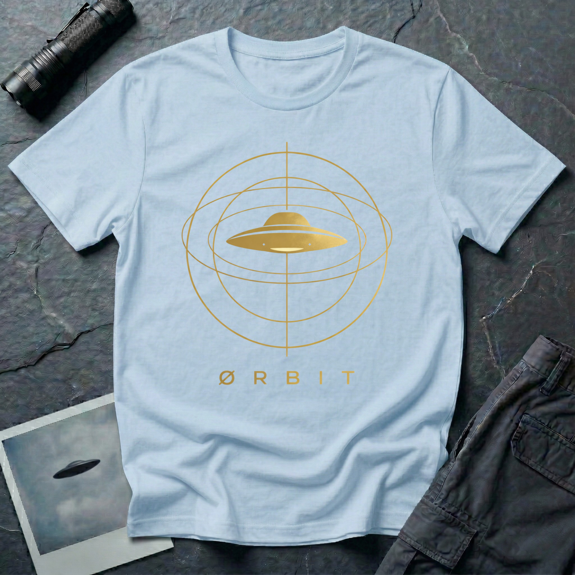 Orbit astronomical UFO t-shirt — flying saucer centred in a gold orbital sphere framework with "Ørbit" sans serif typography on light blue cotton tee