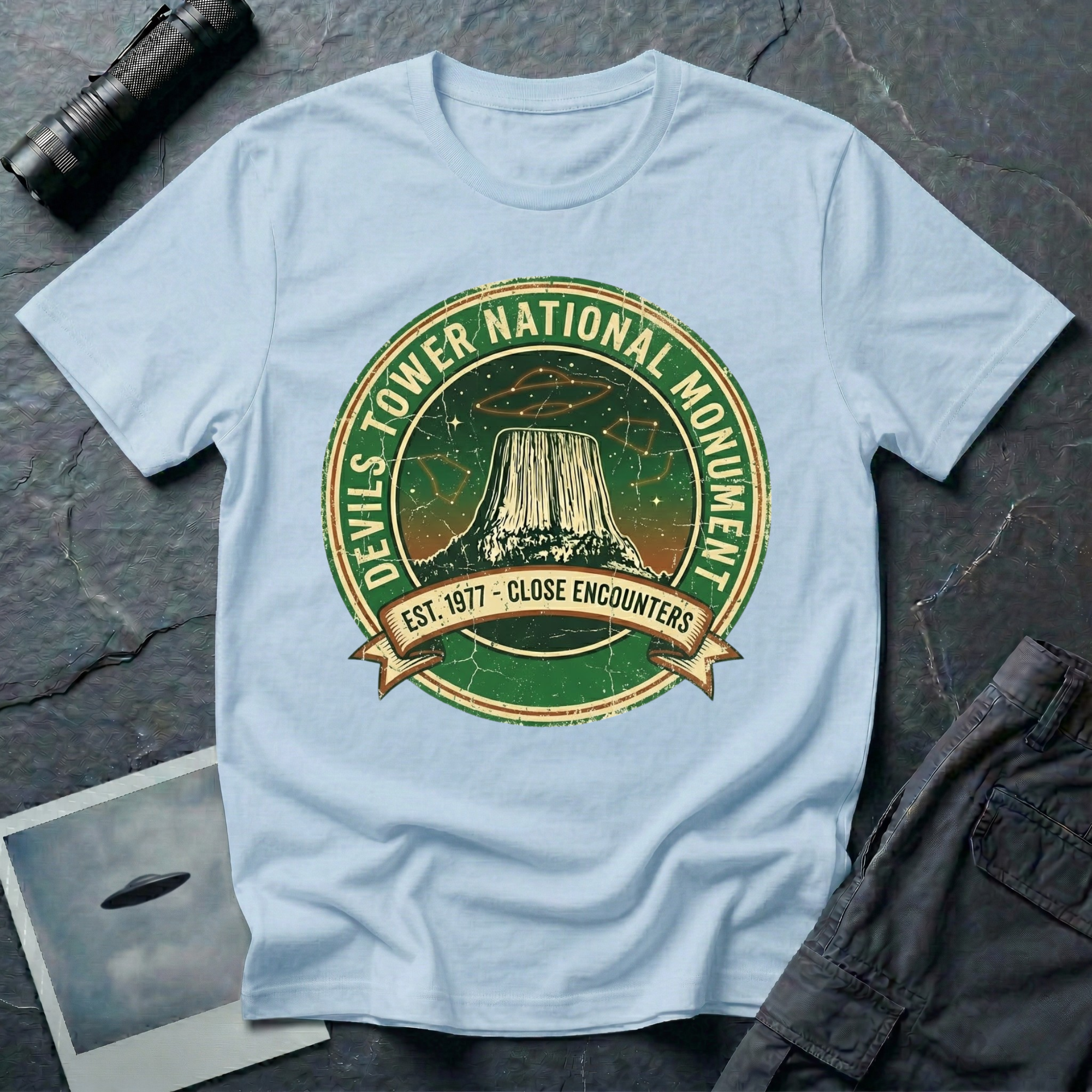 Devils Tower UFO t-shirt — vintage distressed National Park badge with the Wyoming monument silhouette, tiny flying saucers and constellations over a starry green sky, "DEVILS TOWER NATIONAL MONUMENT" arc text and "EST. 1906 — CLOSE ENCOUNTERS" banner on light blue cotton tee