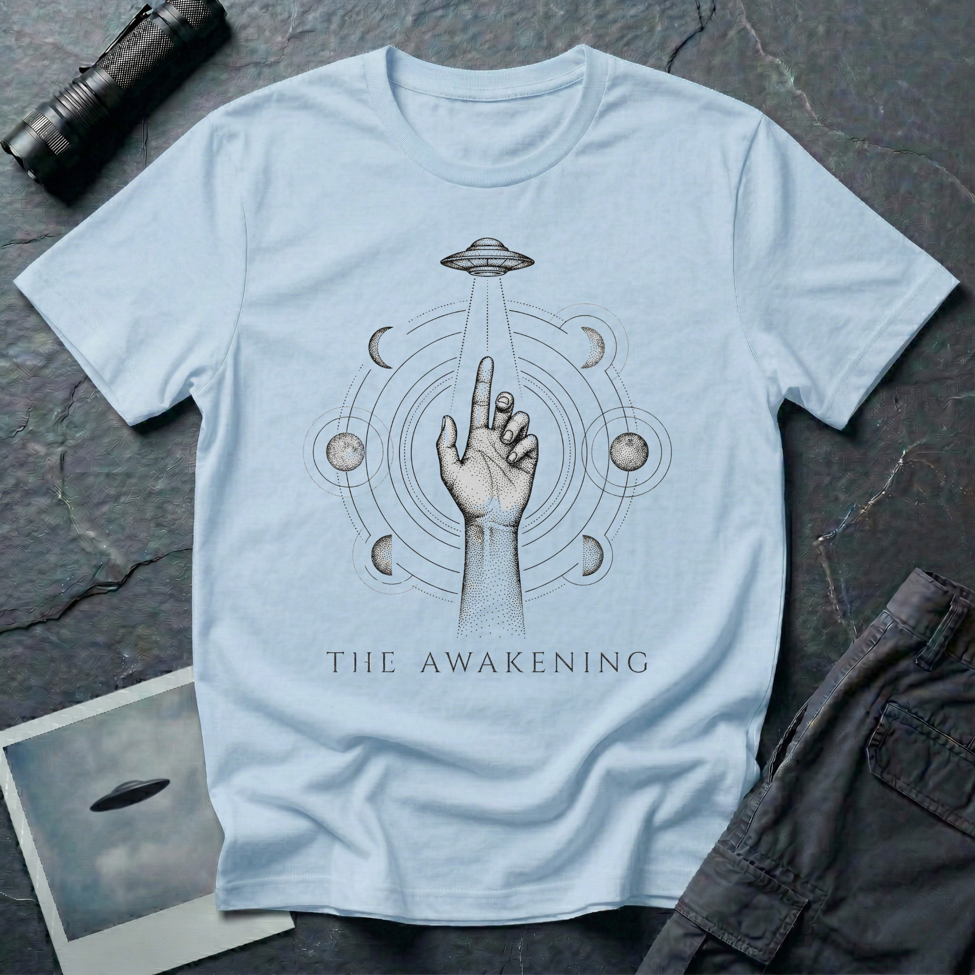 The Awakening mystical UFO t-shirt — hand reaching toward flying saucer beam surrounded by six moon phases in stippled black dotwork on light blue cotton tee