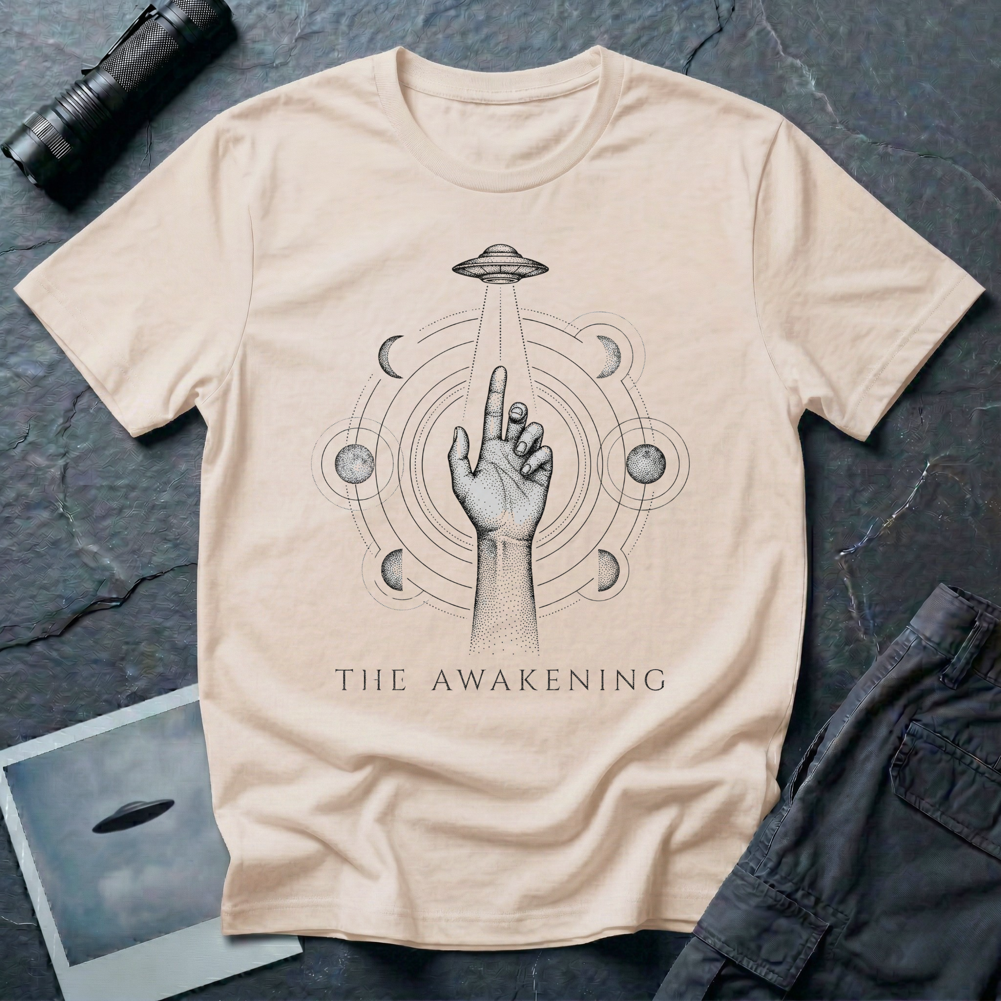 The Awakening mystical UFO t-shirt — hand reaching toward flying saucer beam surrounded by six moon phases in stippled black dotwork on natural cotton tee