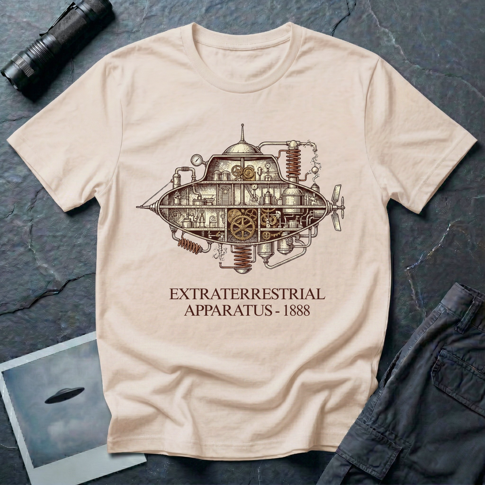 Extraterrestrial Apparatus steampunk UFO t-shirt — Victorian-era airship with exposed brass machinery gears and copper piping with "Extraterrestrial Apparatus 1888" caption on natural cotton tee