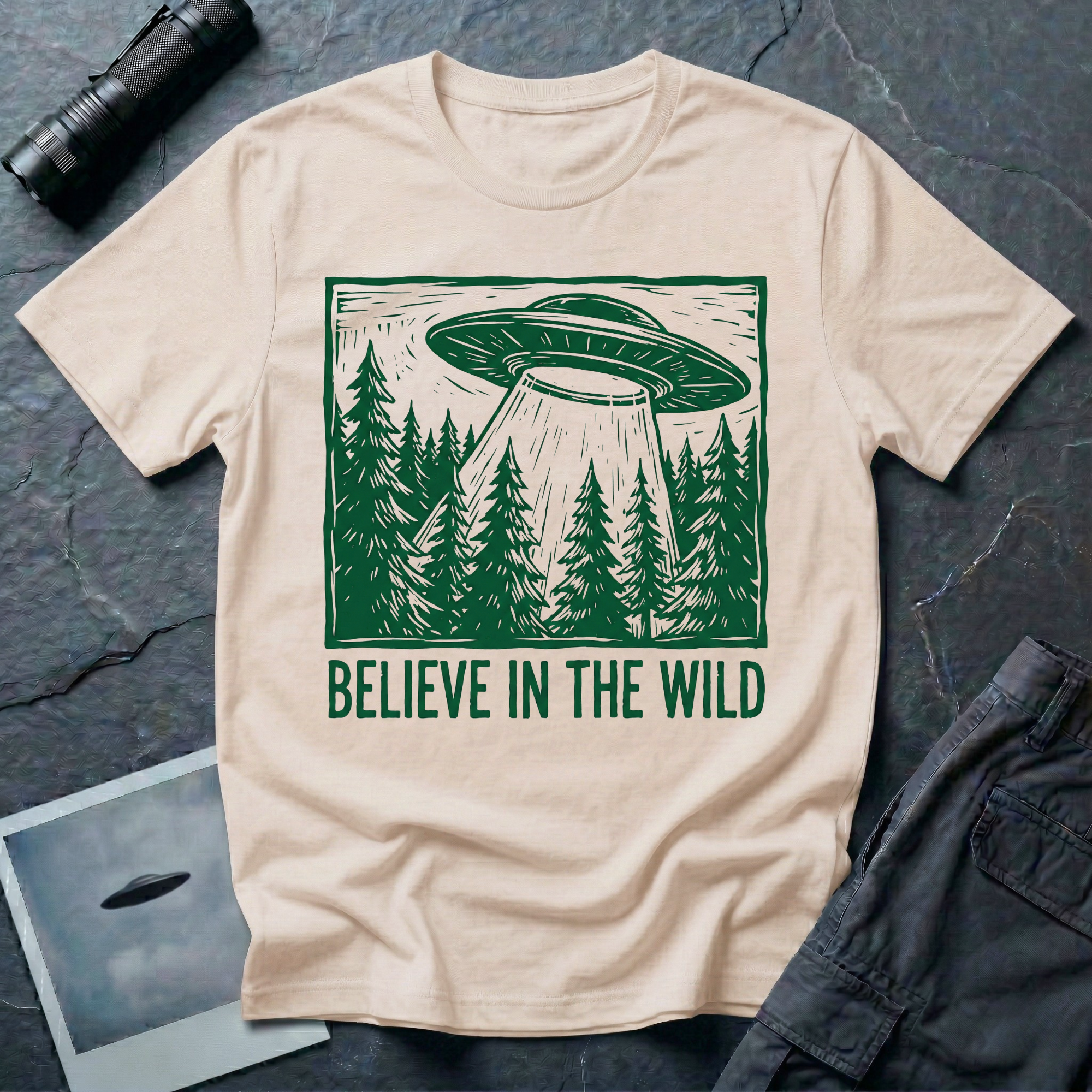 Believe In The Wild forest UFO t-shirt — flying saucer over dense pine forest rendered in green woodcut linocut carved-block print style with "BELIEVE IN THE WILD" caption on natural cotton tee
