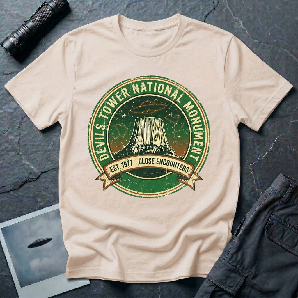 Devils Tower UFO t-shirt — vintage distressed National Park badge with the Wyoming monument silhouette, tiny flying saucers and constellations over a starry green sky, "DEVILS TOWER NATIONAL MONUMENT" arc text and "EST. 1906 — CLOSE ENCOUNTERS" banner on natural cotton tee