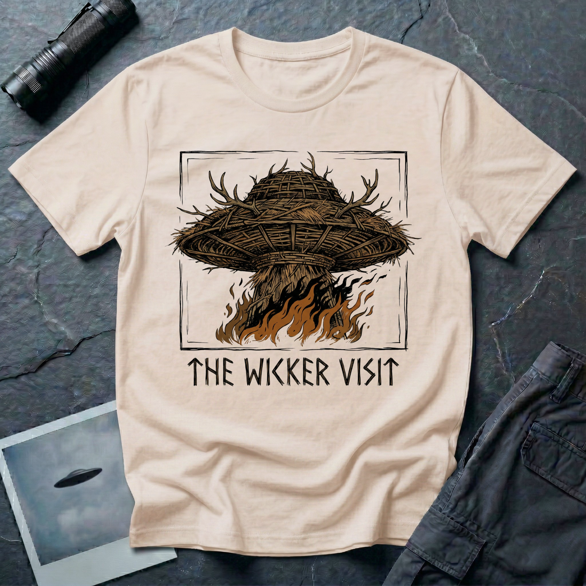 The Wicker Visit folk horror UFO t-shirt — woven wicker flying saucer with antler crown burning in orange flames on natural cotton tee