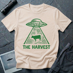 The Harvest dark folklore UFO t-shirt — linocut flying saucer with swirling beam abducting a lone cow in green on natural cotton tee