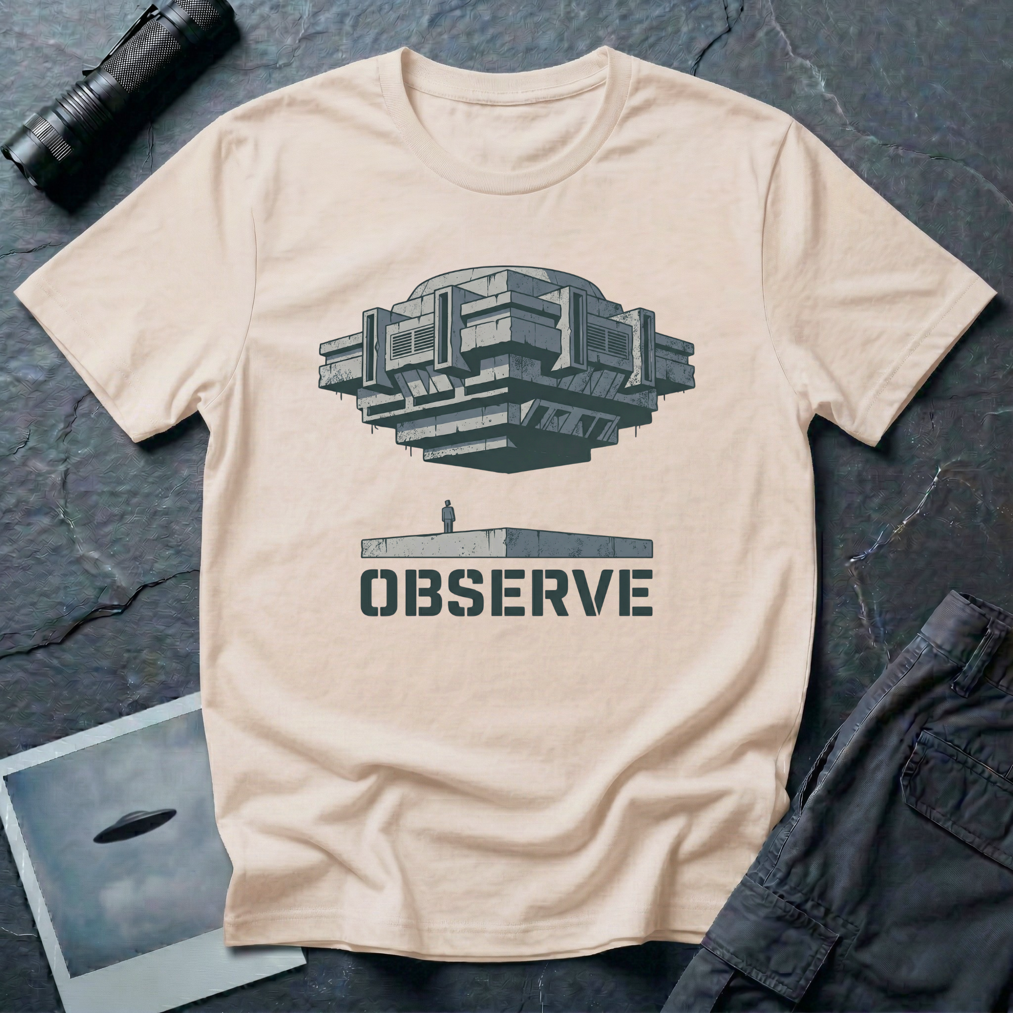 Observe Brutalist UFO t-shirt — massive concrete architectural craft hovering over small figure on plinth with "Observe" stencil type on natural cotton tee