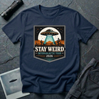 Stay Weird desert UFO t-shirt — tour-poster badge with flying saucer over desert landscape and cacti on navy cotton tee