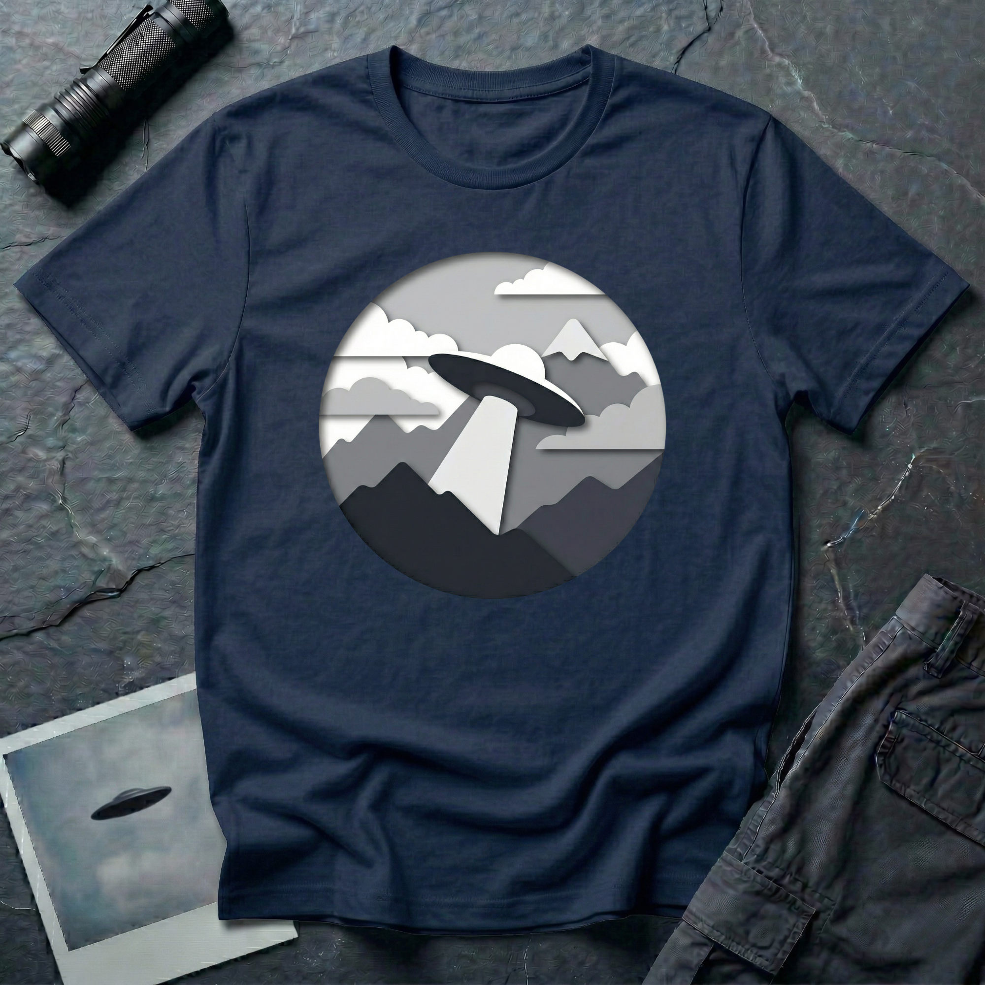 Papercut UFO t-shirt — layered paper-craft illustration of flying saucer over mountains and clouds in grey monochrome on navy cotton tee