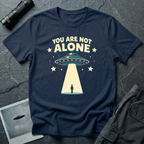 You Are Not Alone friendly UFO t-shirt — retro saucer beaming down on a calm human figure with "You Are Not Alone" text on navy cotton tee