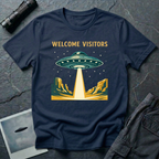Welcome Visitors Southwest UFO t-shirt — flying saucer beaming down into gold and teal desert canyon landscape with "Welcome Visitors" text on navy cotton tee