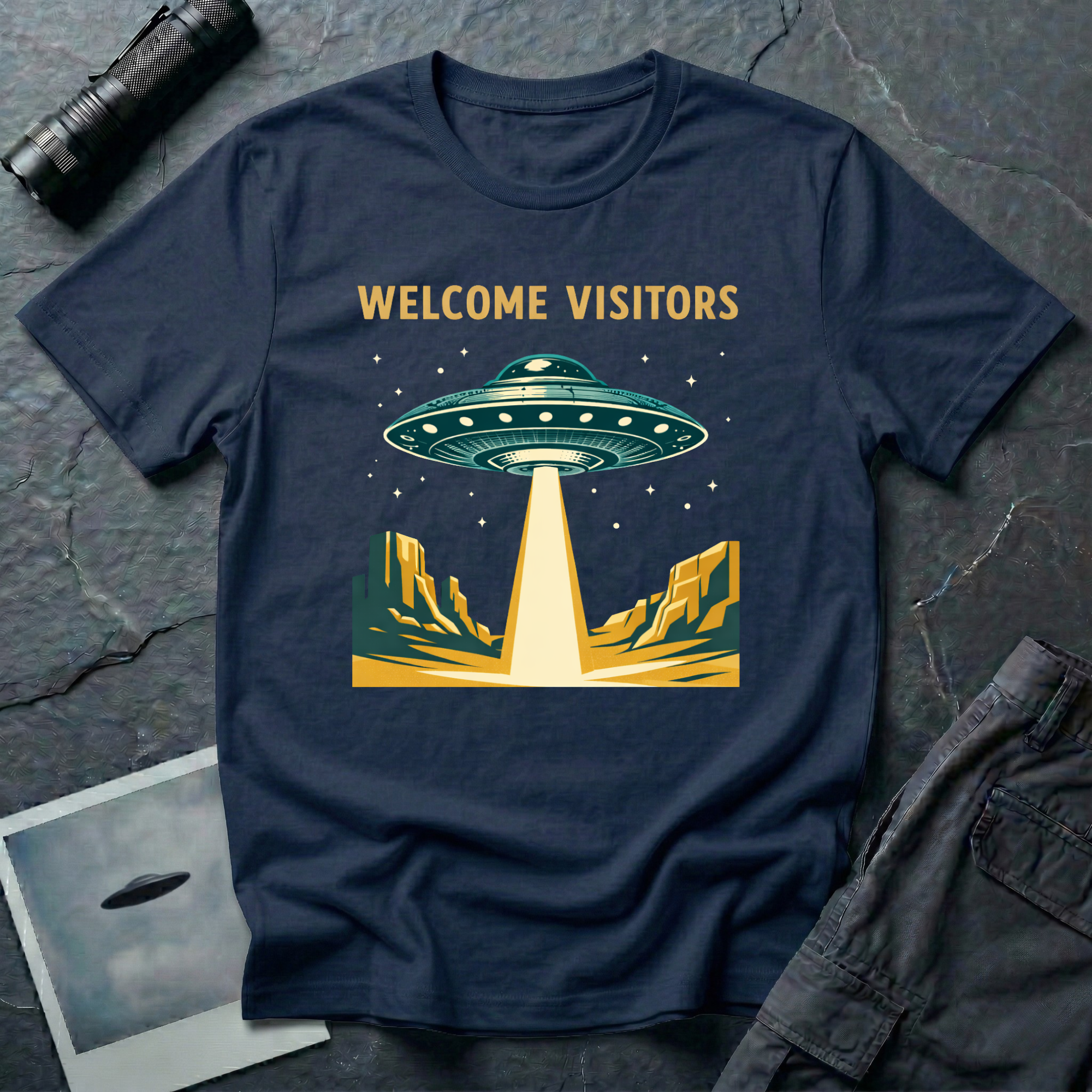 Welcome Visitors Southwest UFO t-shirt — flying saucer beaming down into gold and teal desert canyon landscape with "Welcome Visitors" text on navy cotton tee