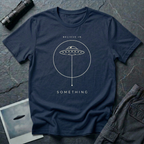Believe in Something minimalist UFO t-shirt — flying saucer inside a circle with a suspended plumb line in cream on navy cotton tee