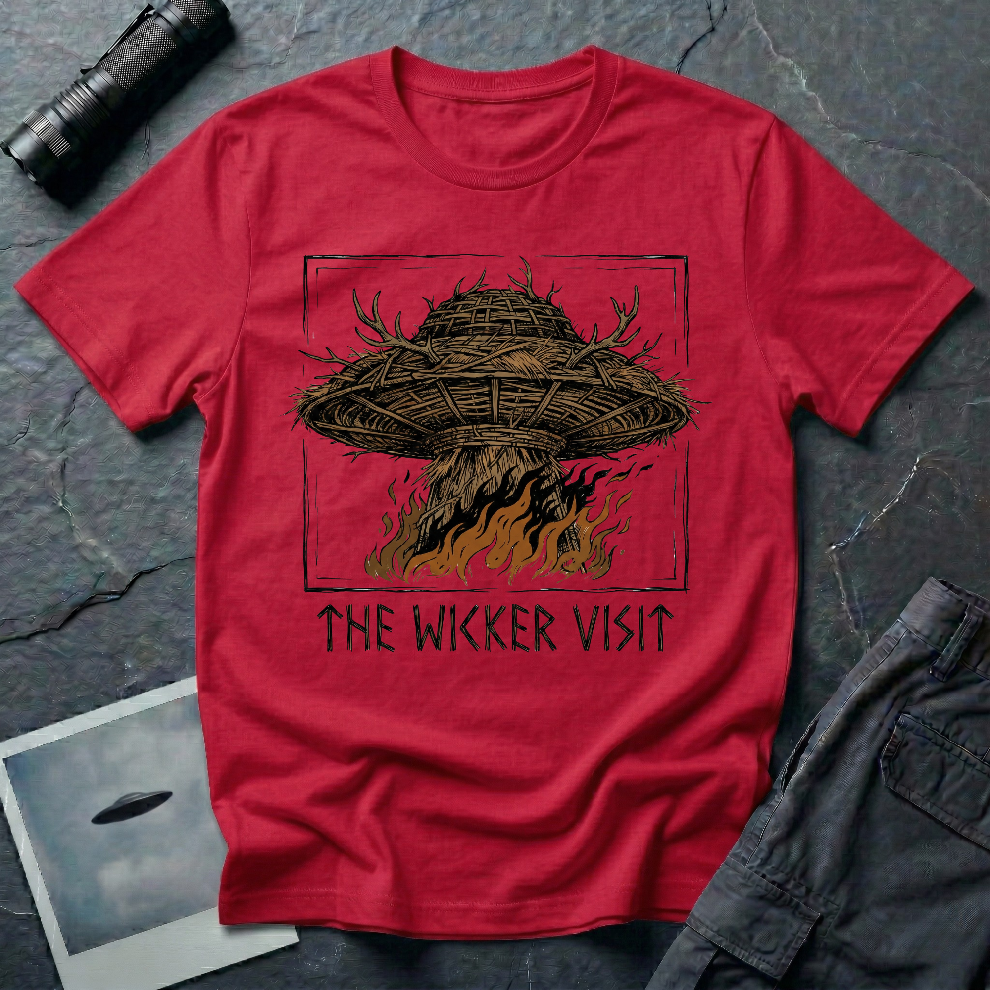 The Wicker Visit folk horror UFO t-shirt — woven wicker flying saucer with antler crown burning in orange flames on red cotton tee