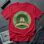 Devils Tower UFO t-shirt — vintage distressed National Park badge with the Wyoming monument silhouette, tiny flying saucers and constellations over a starry green sky, "DEVILS TOWER NATIONAL MONUMENT" arc text and "EST. 1906 — CLOSE ENCOUNTERS" banner on red cotton tee