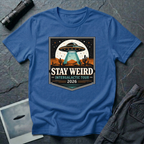 Stay Weird desert UFO t-shirt — tour-poster badge with flying saucer over desert landscape and cacti on blue cotton tee