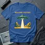 Welcome Visitors Southwest UFO t-shirt — flying saucer beaming down into gold and teal desert canyon landscape with "Welcome Visitors" text on blue cotton tee