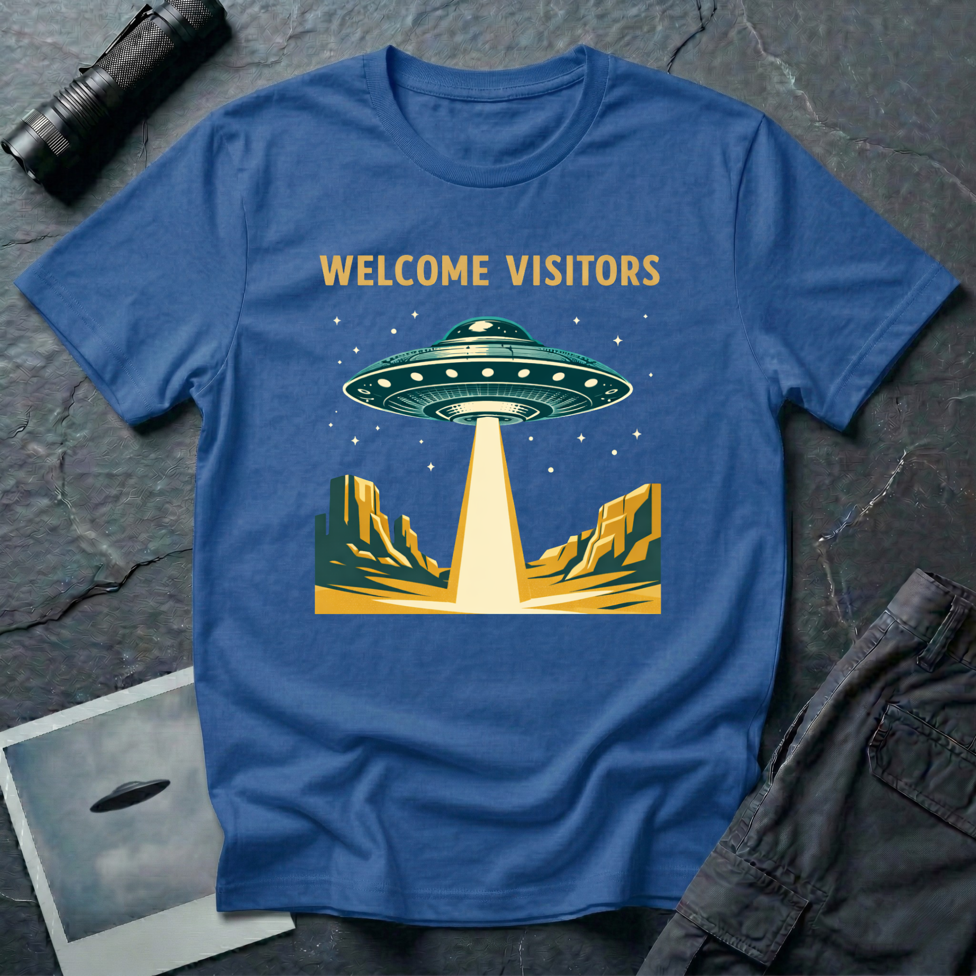 Welcome Visitors Southwest UFO t-shirt — flying saucer beaming down into gold and teal desert canyon landscape with "Welcome Visitors" text on blue cotton tee