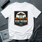 Stay Weird desert UFO t-shirt — tour-poster badge with flying saucer over desert landscape and cacti on white cotton tee