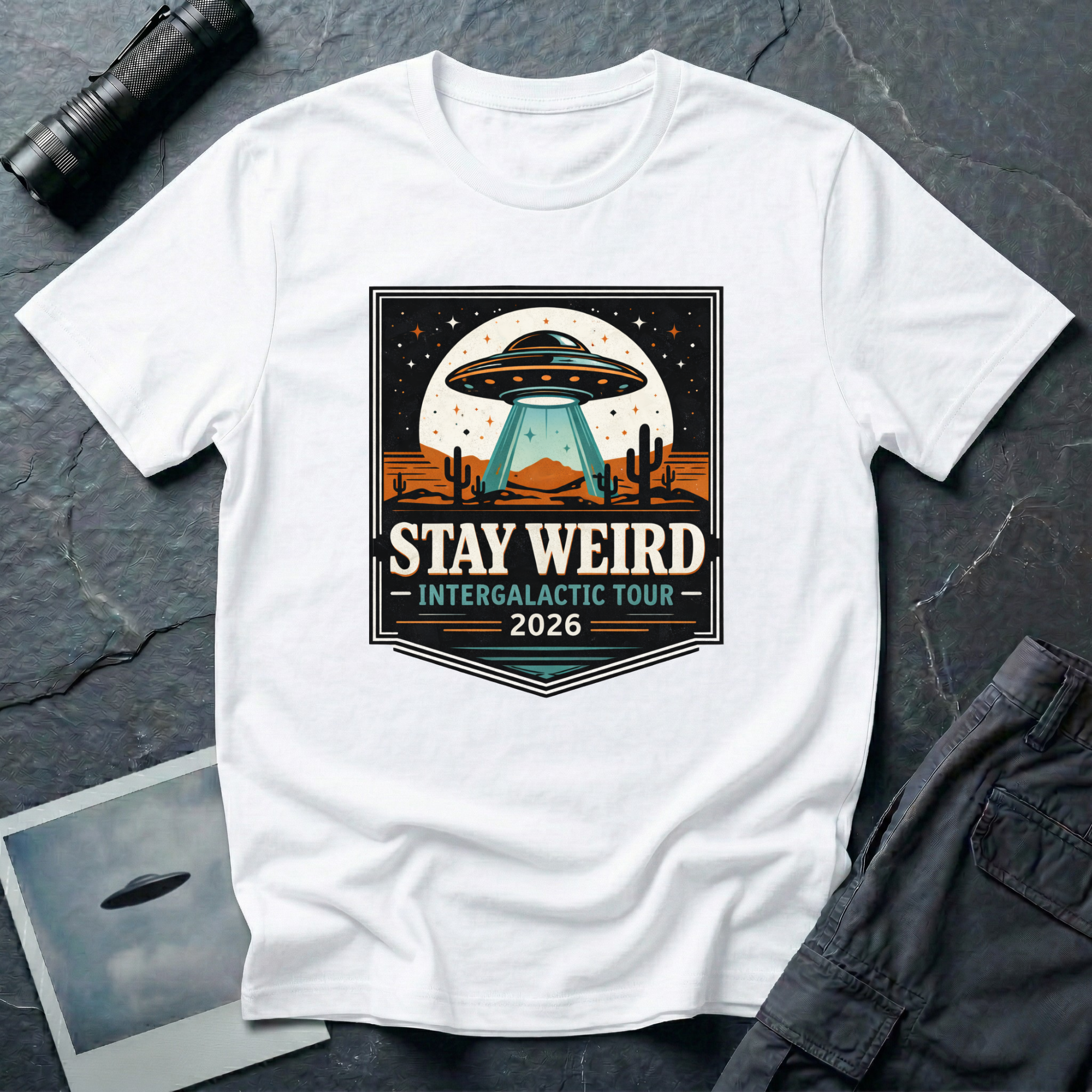 Stay Weird desert UFO t-shirt — tour-poster badge with flying saucer over desert landscape and cacti on white cotton tee